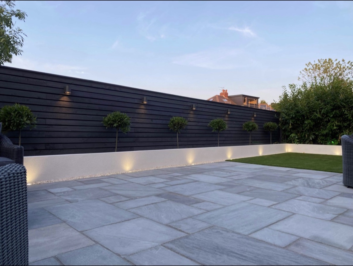 A finished garden at dusk: sandstone paving, a rendered raised bed and lighting set into a dark slatted fence.